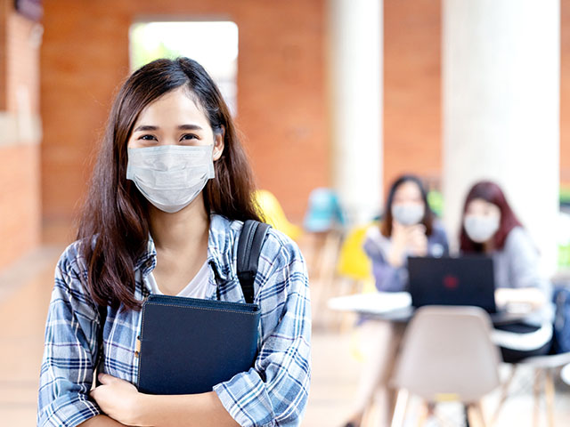 A woman in a face mask standing holding a notebook, with two students using a laptop in the background.